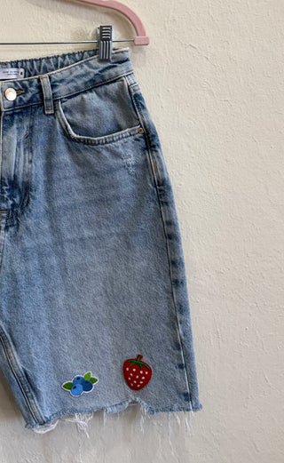 Light wash denim mini skirt with a raw hem, featuring embroidered berry patches on the front, hanging on a pink hanger against a textured off-white wall.