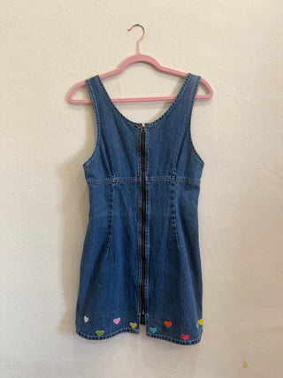 A sleeveless blue denim dress with a front zipper, hanging on a pink hanger against a textured off-white wall. Small colorful heart details are embroidered near the bottom hem.