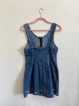 A blue denim overall-style dress with colorful embroidered hearts near the hem, hanging on a pink hanger against a textured off-white wall.