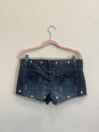 Back view of blue denim shorts with white embroidered flower details, hanging on a pink hanger against a textured off-white wall.
