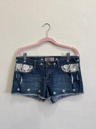 Distressed denim shorts with floral lace appliques on the front pockets and white embroidered star details, hanging on a pink hanger against a textured off-white wall.
