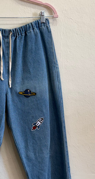 Blue denim drawstring pants hanging on a pink hanger against a textured off-white wall, featuring embroidered space-themed patches including a Saturn-like ringed planet and a small rocket ship on the left leg.