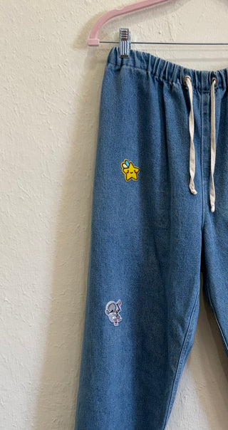 Blue denim drawstring pants hanging on a pink hanger against a textured off-white wall, featuring a yellow star patch on the thigh and a small white embroidered patch near the lower leg.