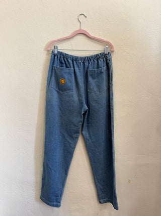 A pair of blue denim pants with an elastic waistband and a small yellow smiley face patch on the back pocket, hanging on a pink hanger against a textured beige wall.