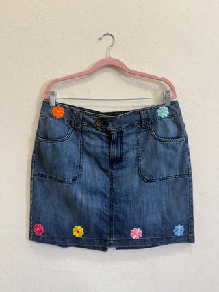 A blue denim mini skirt with colorful embroidered flowers, hanging on a pink hanger against a textured off-white wall.