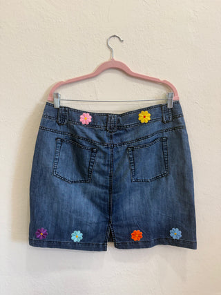 A pair of blue denim shorts hanging on a pink hanger against a textured off-white wall. The shorts feature colorful embroidered flower details in pink, yellow, purple, blue, and orange.