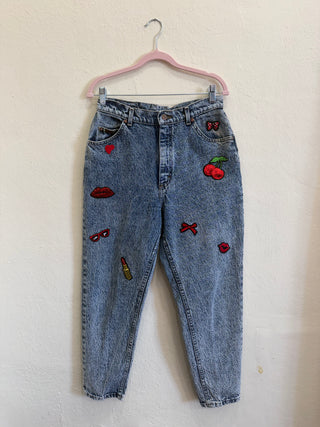 Light wash blue denim jeans with colorful embroidered accessories including cherries, lipstick, sunglasses, and a heart, hanging on a pink hanger against a textured off-white wall.