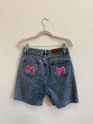 A pair of blue denim shorts hanging on a pink hanger against a textured off-white wall. The back of the shorts features two pink bow patches on the back pockets.
