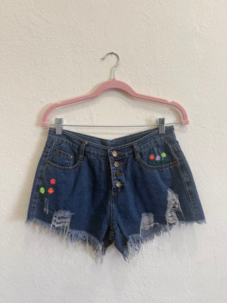 Distressed denim shorts with a button fly and colorful embroidered flowers, hanging on a pink hanger against a textured white wall.