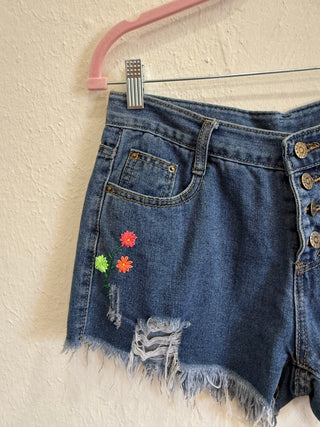 A pair of distressed denim shorts with colorful embroidered flowers on the left leg, hanging from a pink hanger against a textured beige wall.