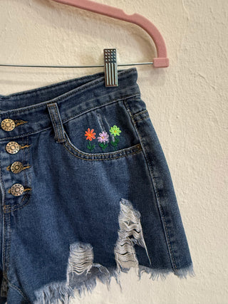 Close-up of a pair of distressed denim shorts hanging on a pink hanger against a textured cream wall. The shorts feature colorful embroidered flowers on the pocket, decorative gold-tone buttons on the waistband, and frayed raw hems.