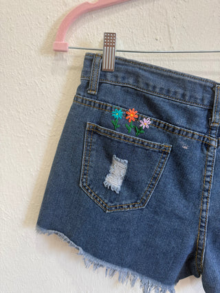 Close-up of the back of a pair of blue denim shorts hanging on a pink hanger against a textured off-white wall. The shorts have a distressed ripped pocket with white frayed denim inside, colorful embroidered flowers on the back pocket, and a frayed hem.