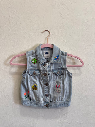 A light blue denim vest with embroidered bunny, bee, and flower patches, hanging on a pink hanger against a textured off-white wall.