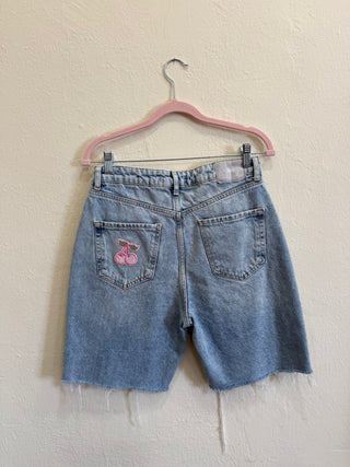 Light wash blue denim shorts with a pink cherry embroidery on the back pocket, hanging on a pink hanger against a textured beige wall.