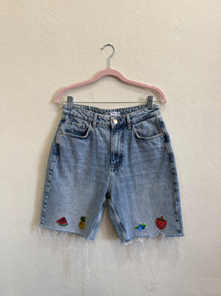 Light wash denim shorts with a raw frayed hem and colorful embroidered fruit patches, hanging on a pink hanger against a textured off-white wall.